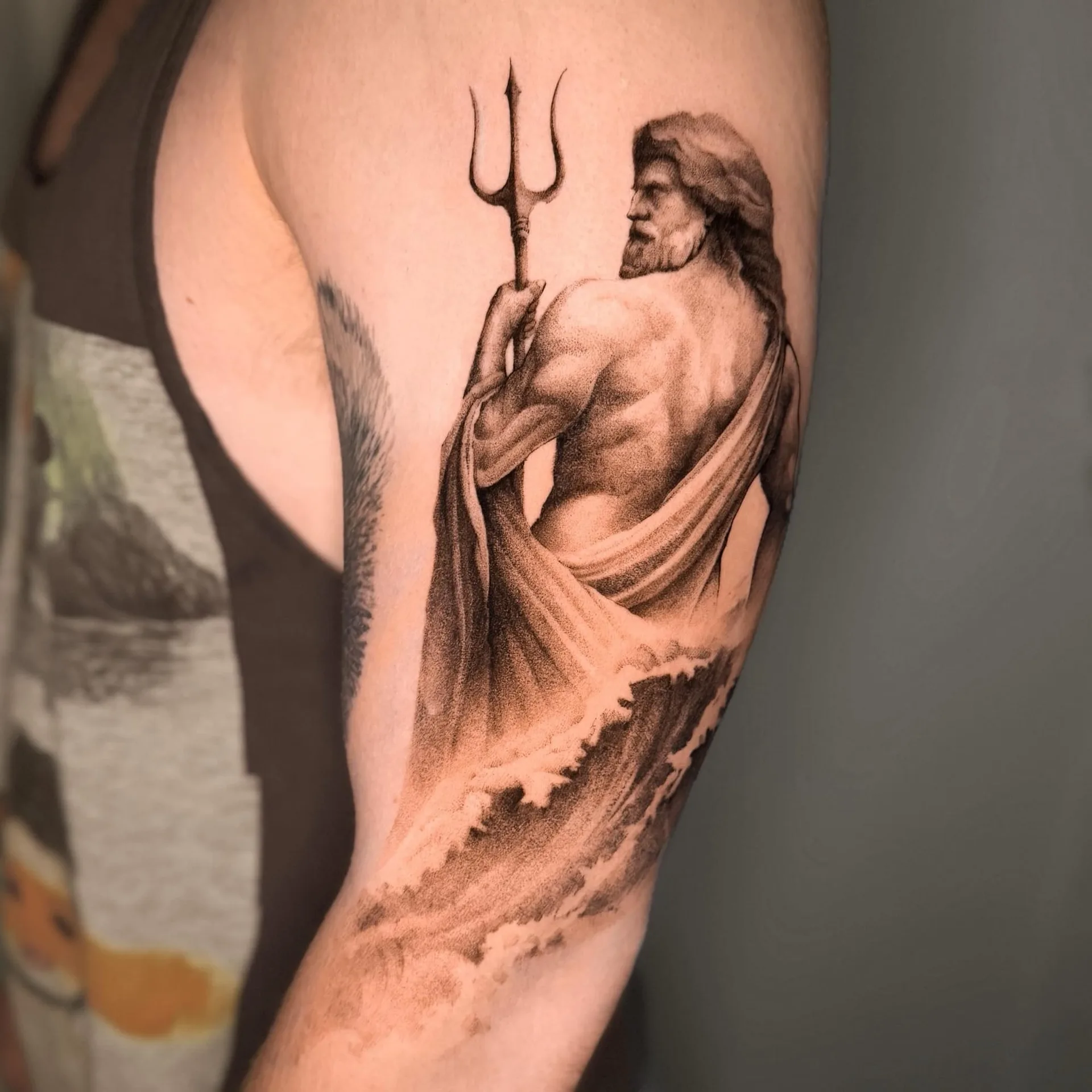 Poseidon mit Dreizack und wehendem Tuch, darunter Wellen. Black-and-Grey-Tattoo von Inna auf dem Oberarm.