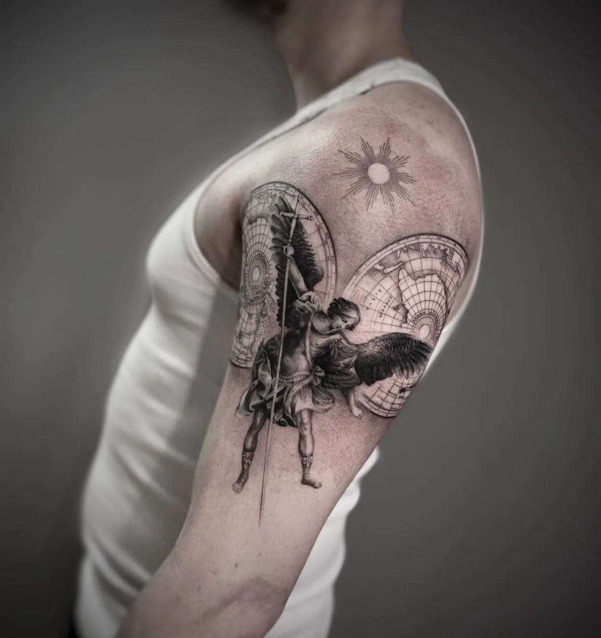 Erzengel mit Schwert und Flügeln vor einer alten Weltkarte, darüber eine Sonne. Black-and-Grey-Tattoo von Sloan auf dem Oberarm.