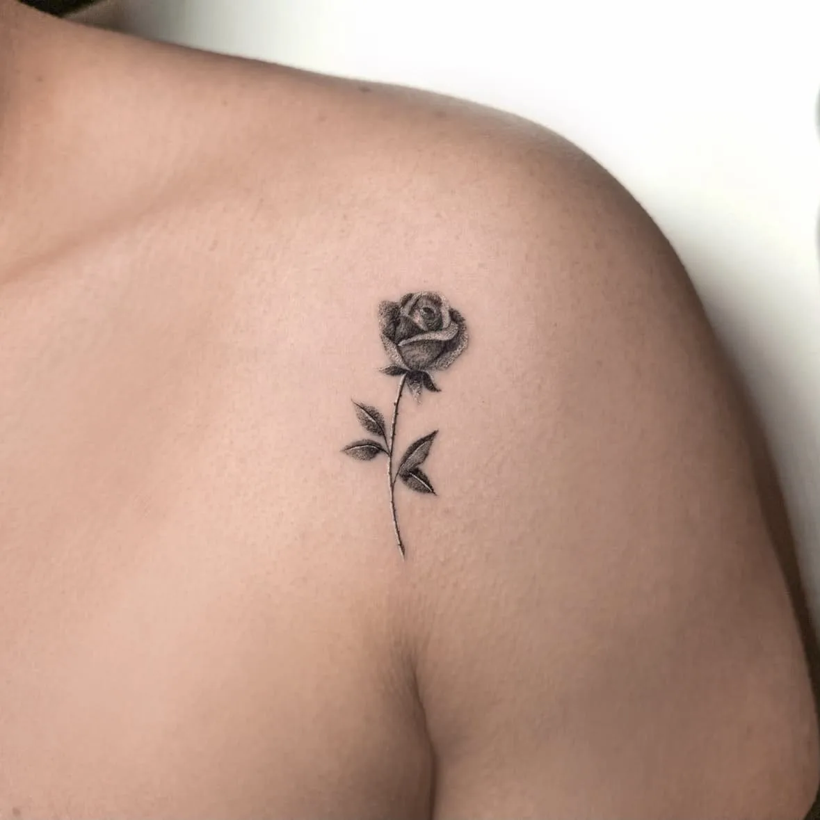 Rose mit Stiel und zwei Blättern, fein schattiert. Fine-Line-Tattoo von Inna an der Schulter.