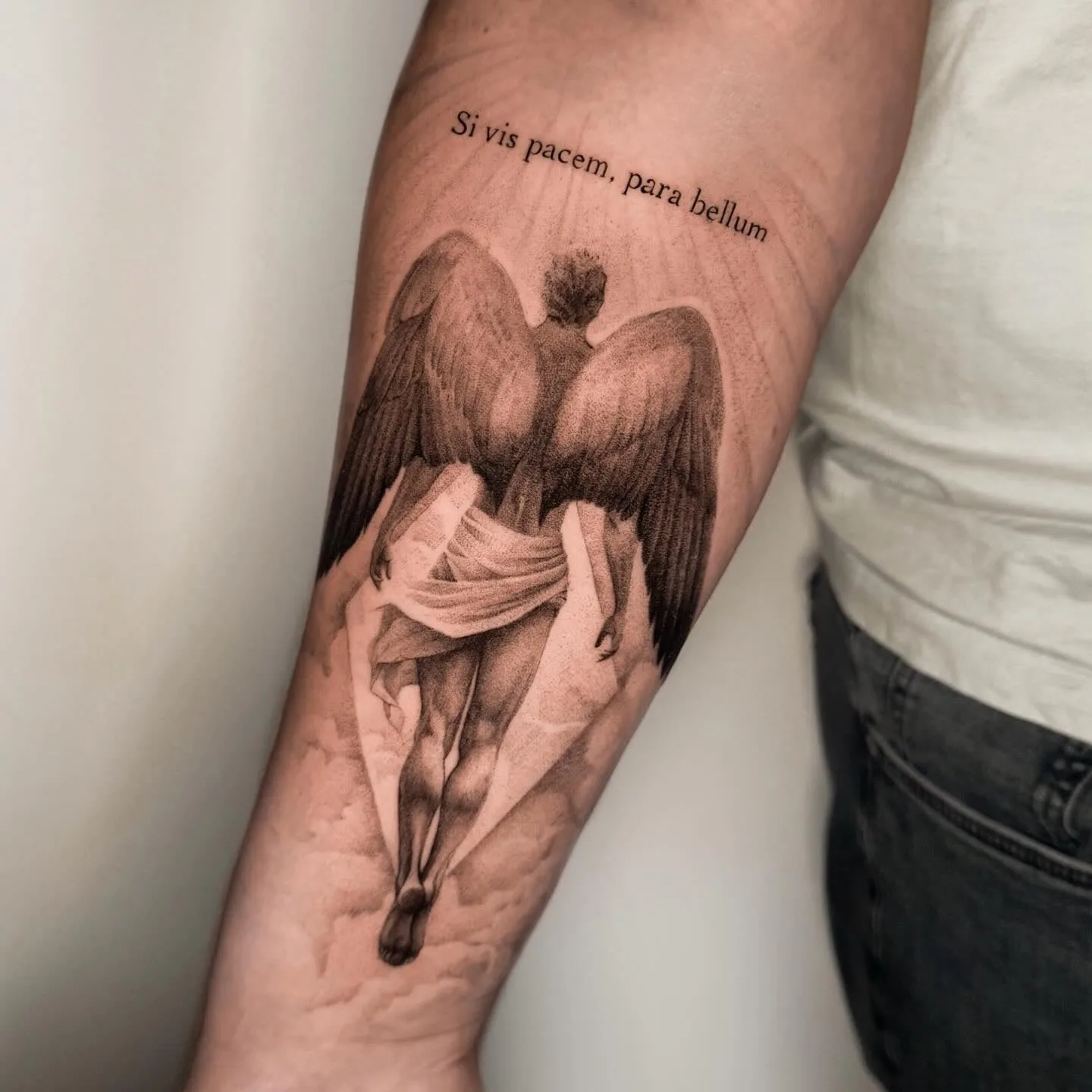 Engel von hinten mit großen Flügeln, der in die Wolken steigt, darüber ein lateinischer Satz. Black-and-Grey-Tattoo von Inna auf dem Unterarm.