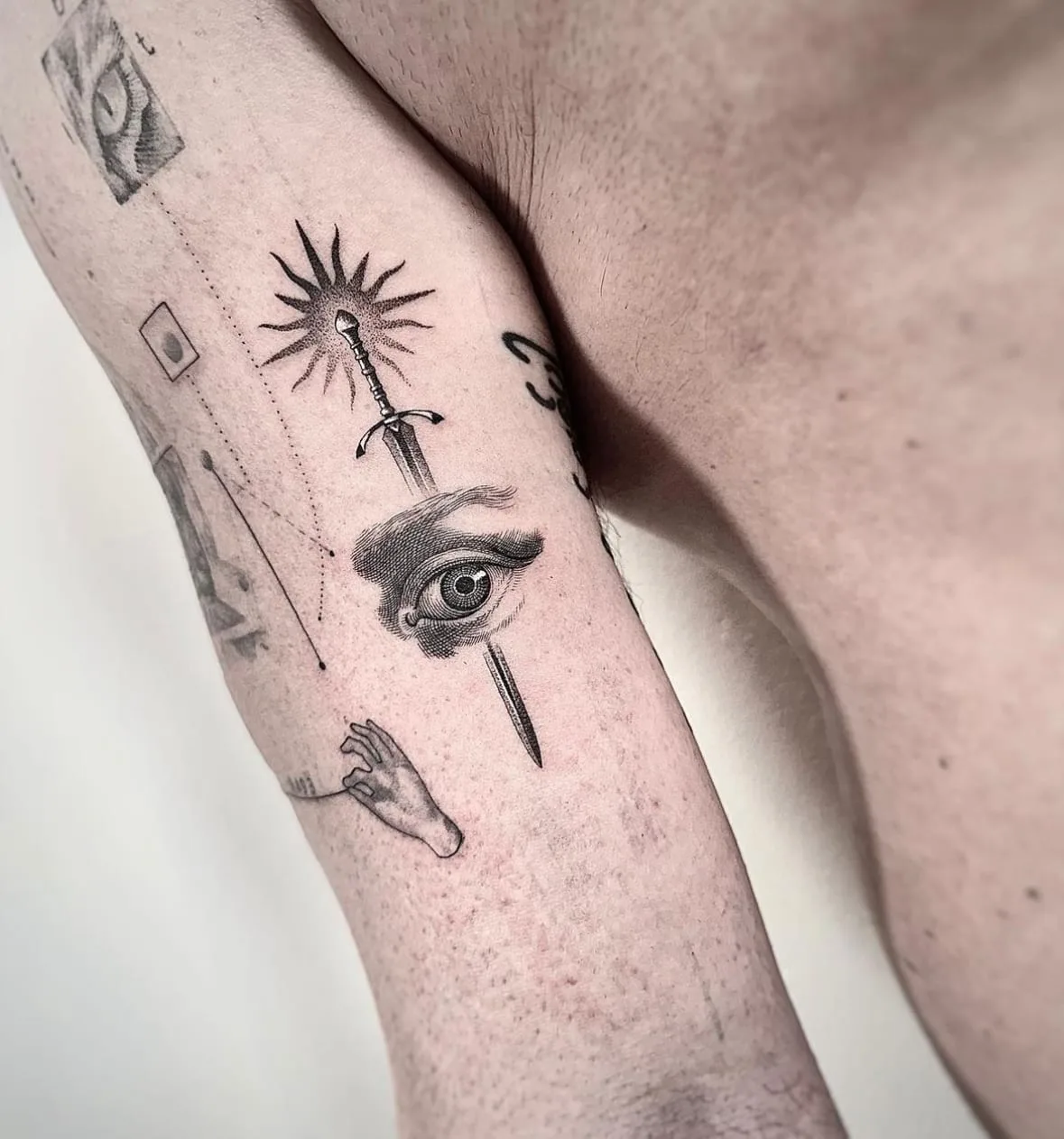 Schwert mit Strahlenkranz, das durch ein Auge führt, darunter eine Hand. Black-and-Grey-Tattoo von Sloan auf dem Oberarm.