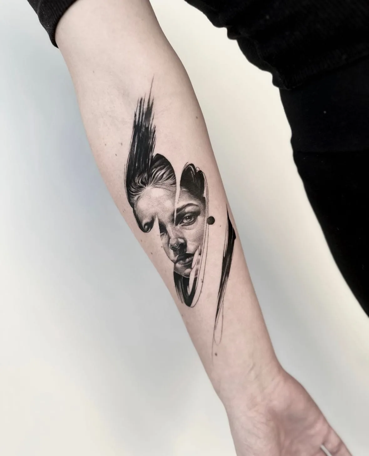 Geteiltes Frauengesicht zwischen schwarzen Pinselstrichen. Black-and-Grey-Tattoo von Sloan auf dem Unterarm.