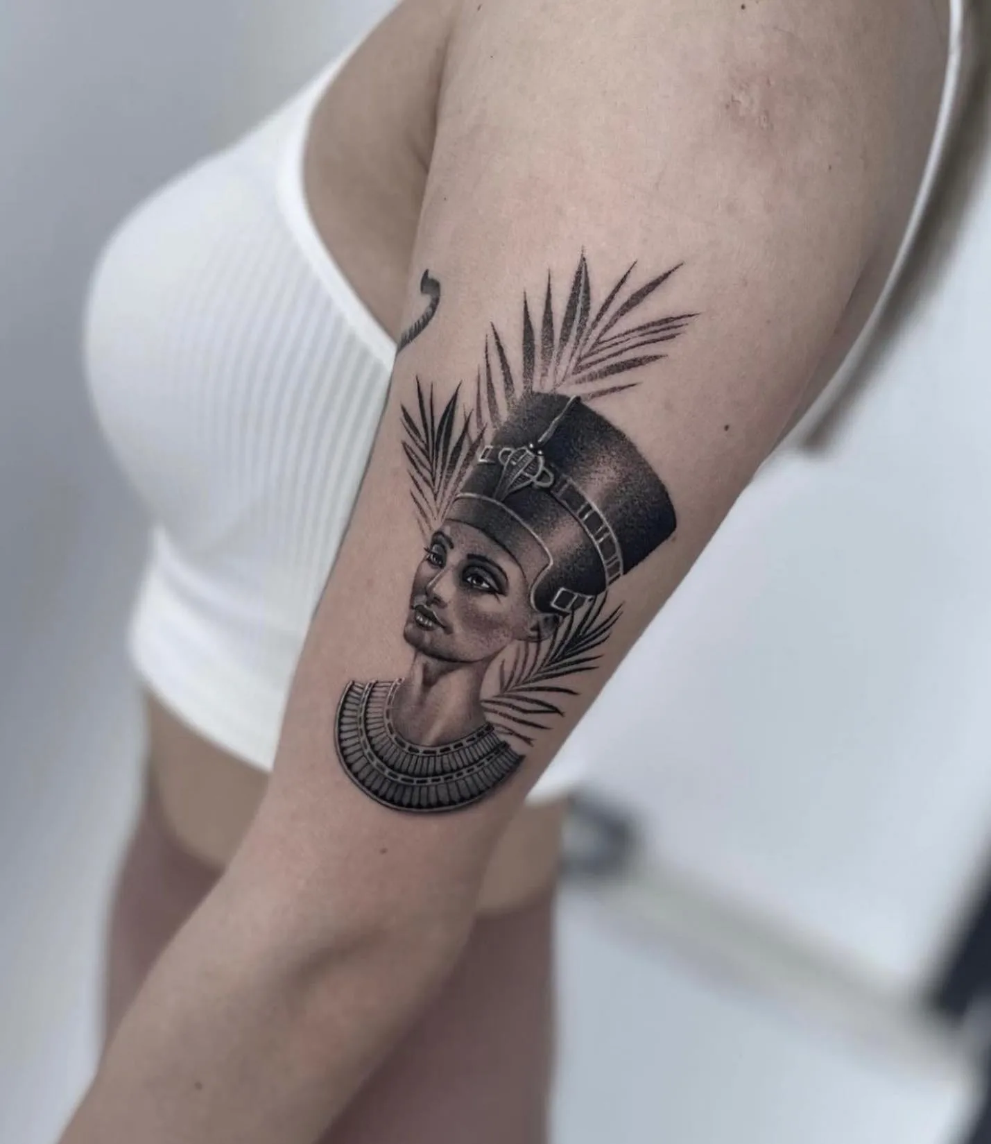 Büste der Nofretete mit Krone und Halskragen vor Palmblättern. Black-and-Grey-Tattoo von Sloan auf dem Oberarm.