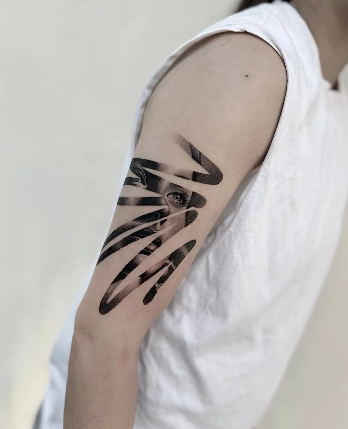 Augen und Lippen eines Frauengesichts zwischen geschwungenen schwarzen Streifen. Black-and-Grey-Tattoo von Sloan auf dem Oberarm.