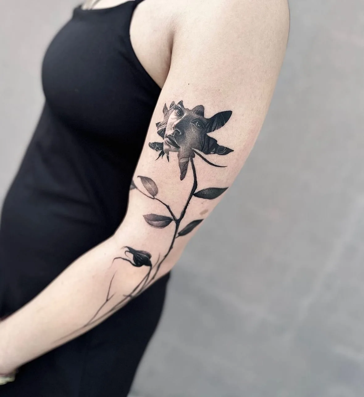 Blüte mit einem Frauengesicht, der Stiel mit Blättern läuft den Arm hinab. Black-and-Grey-Tattoo von Sloan auf dem Oberarm.