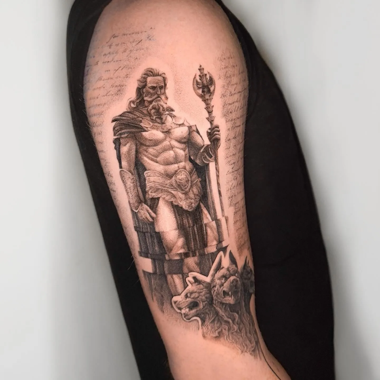 Hades mit Zepter vor einer Säule, zu seinen Füßen Kerberos, daneben Schrift. Black-and-Grey-Tattoo von Inna auf dem Oberarm.