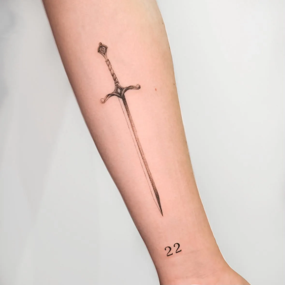 Schwert mit verziertem Griff, darunter die Zahl 22. Fine-Line-Tattoo von Inna auf dem Unterarm.