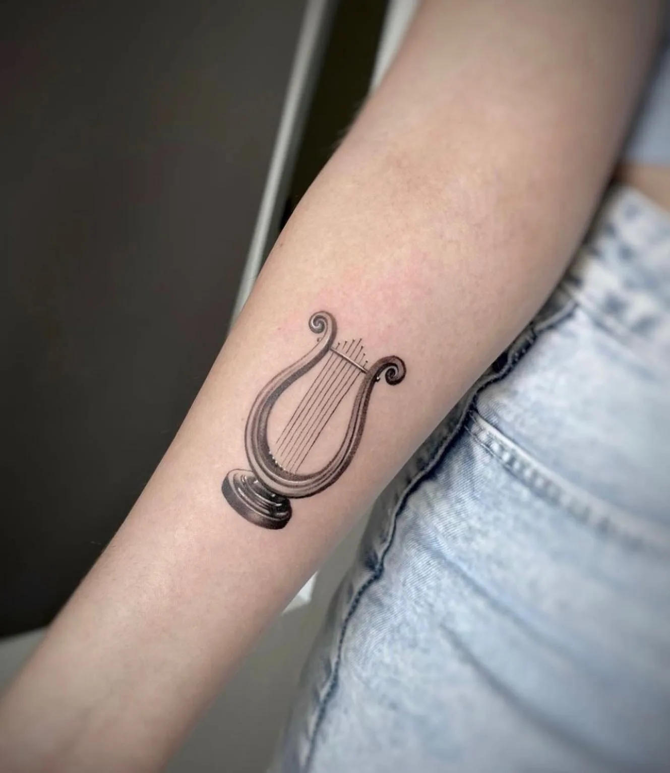 Kleine Lyra mit feinen Saiten auf einem Sockel. Black-and-Grey-Tattoo von Sloan auf dem Unterarm.